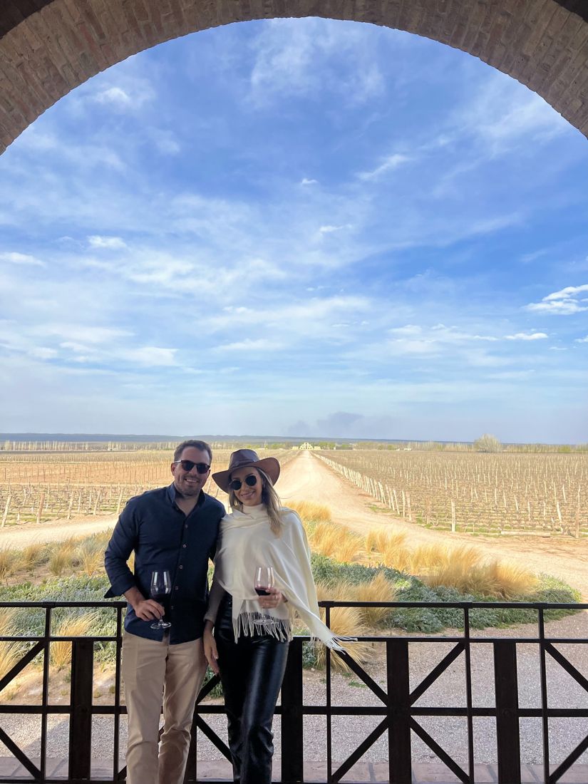 Paula e Francisco na varanda de uma vinícola, sob um arco de tijolos